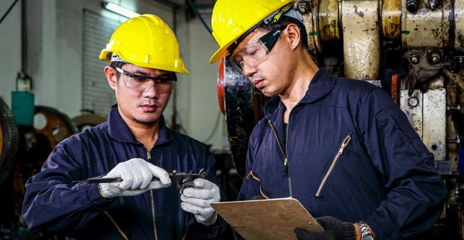 Two Asian male workers in safety clothing use Vernier calipers. Inspect the quality and measure the spare parts of working machines in industrial plants and production processes.