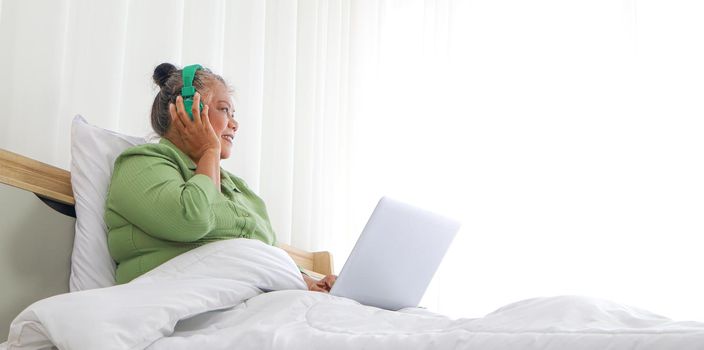 Retirement women Asians, use a laptop and headphones, happy in bed, concept listening to music, and living at home. Long-distance conversation through the online system Use of technology