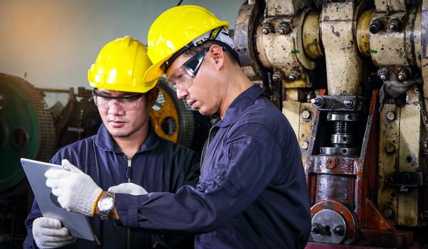 Two skilled Asian workers, dressed in uniforms, safety hats, and safety goggles. Are using a tablet to analyze data Metal press maintenance inspection at the factory, teamwork concept