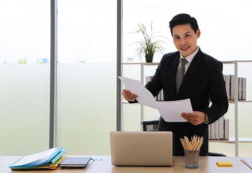 Asian businessman That is positive adults Stand up and hold documents Look at the camera and smile and relax in a modern office. Concepts that work successfully There is a computer on the desk.
