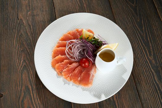From above view of delicious raw salmon slices on white plate served with lemon, onion, tomatoes, sprouts and small jar of oil. Tasty appetizer on wooden table in luxury restaurant for dinner.