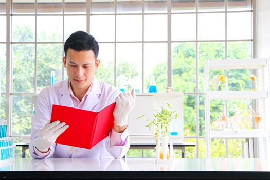 A scientist or researcher in an Asian male wearing a white coat is working and reading a book to find information. Being in a scientific laboratory With test tube equipment