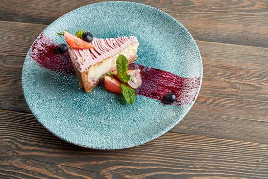 From above view of traditional cheesecake served with blueberries and cranberries on plate with purple brushstroke decoration. Tasty food on wooden table in restaurant. Concept of dessert, serving.