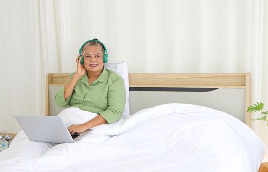 Retirement women Asians, use a laptop and headphones, happy in bed, concept listening to music, and living at home. Long-distance conversation through the online system Use of technology