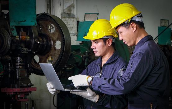 Two skilled Asian workers, dressed in helmet uniforms, are using a computer to analyze the metal press maintenance inspection data. The concept of teamwork at the factory