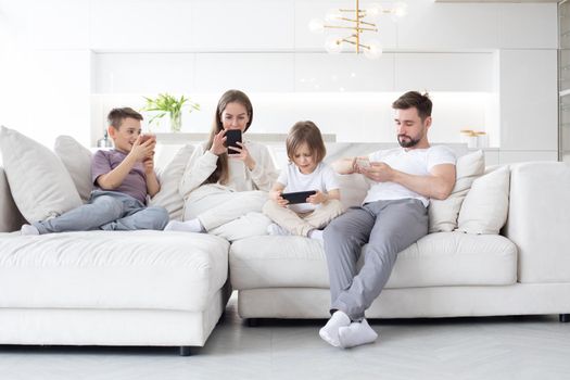 Caucasian parents and kids use devices together sit on sofa, tech addicted family with children hold phone digital tablet having fun with gadgets at home, technology dependence