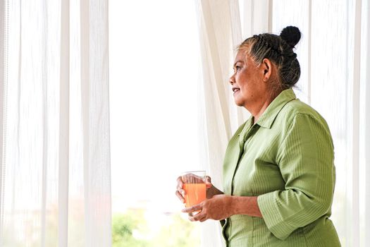 Senior Asian women Drinking orange juice standing by the window in the background of the room, the concept of living the elderly, retirement and healthy lifestyle.