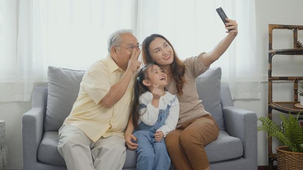 Happy family time relax. Asian grandparents laughing taking selfie with granddaughter on sofa at home by mobile smartphone. senior man or grandpa, daughter and girl kid in living room together