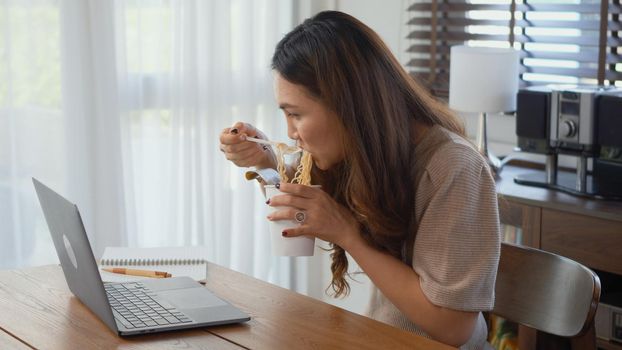 Asian business woman eating instant noodles while working on laptop computer at home office, Happy beautiful young female sitting on desk work overtime doing deadline project, late time business