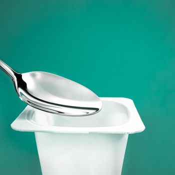 Yogurt cup and silver spoon on green background, white plastic container with yoghurt cream, fresh dairy product for healthy diet and nutrition balance.