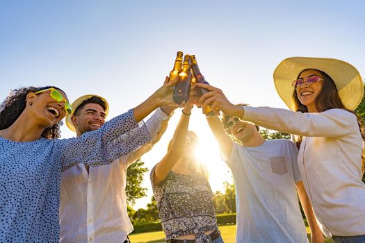 Group of young hipster friends celebrating outdoor drinking beer and toasting with bottle in front of sun setting. Happy beautiful millennials having fun with alcohol outdoor in a park at dusk