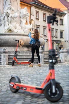 Traveler exploring old town on environmentally friendly electric scooter. Female tourist exploring Ljubljana's old medieval historical city center, taking picture of a fountain with her phone