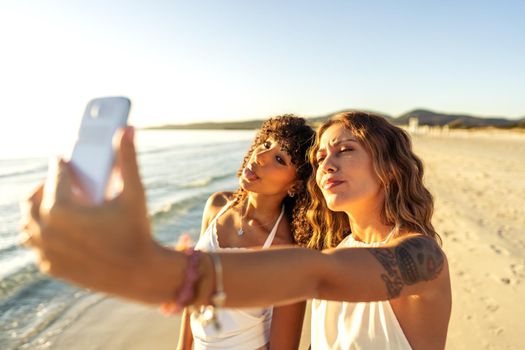 Two cute girls in boho white dress in summer vacation using smartphone taking a selfie making facves standing on sea sand of a tropical beach. Multiracial women travellers having fun with technology