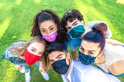 Bright vivid color effect photography of multiracial group of young people looking at camera from bottom wearing colored protective face mask on a green field. New norla outdoor activity limitations