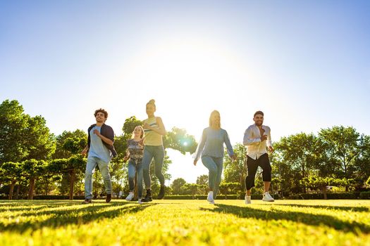 Suggestive low angle view of group of young multiracial gen-z friends running on green field with setting sun at their back with light effect and long shadows on grass. Concept of people team success