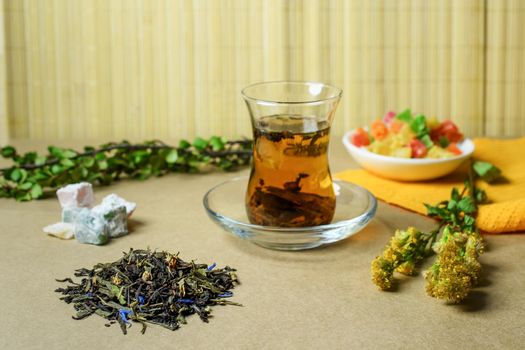 The Turkish traditional glass with tea on a table, nearby a small group of the filled dry tea, the Turkish sweets and a grass