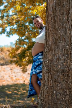 Photo of a young couple behind a tree trunk. Only the young man's head and the pregnant young woman's belly are visible, as well as her feet.