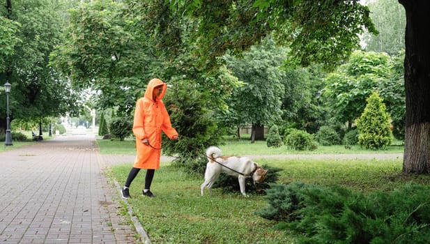 Rainy weather. Young woman in orange raincoat walking with her dog in a summer park