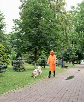 Rainy weather. Young woman in orange raincoat walking with her dog in a summer park