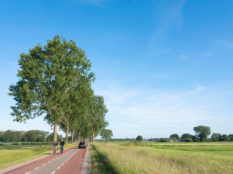 nijmegen, netherlands, 16 june 2021: couple rides bicycle on country road with high trees near nijmegen in holland under blue summer sky