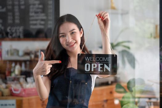 Portrait of smiling young barista girl in apron holding open sign board while standing at her cafe. elegant asian coffee shop female staff turn door plate in the morning in own store small business.