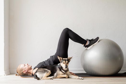 Sport and healthy lifestyle concept. Young sportive woman working out on the fitness ball at home, dog sleeping nearby