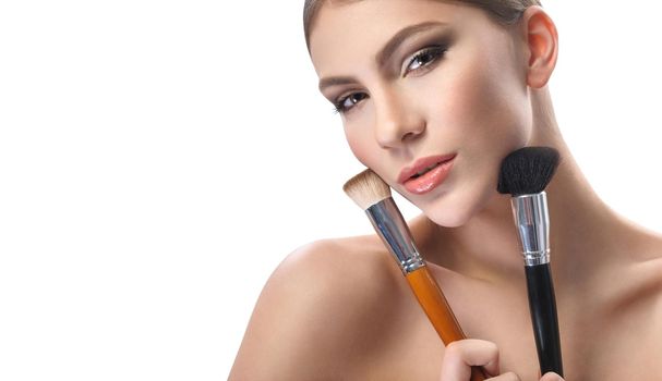 Beauty must haves. Horizontal shot of a beautiful young woman looking to the camera confidently holding makeup brushes to her face wearing professional makeup with brown smoky eyes and nude lipstick