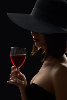 Red wine. Vertical portrait of a mysterious woman in a hat hiding her face having a glass of red wine in a dark room with mysterious lighting incognito secret night evening luxury winery concept