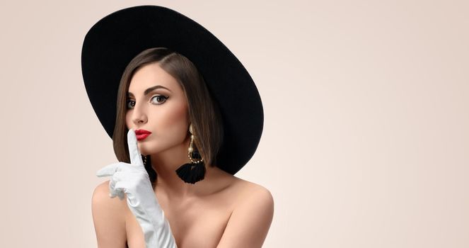 Horizontal studio portrait of a beautiful fashion model wearing a hat and gloves making shushing gesture to the camera copyspace beauty cosmetology skincare secrecy silence concept .
