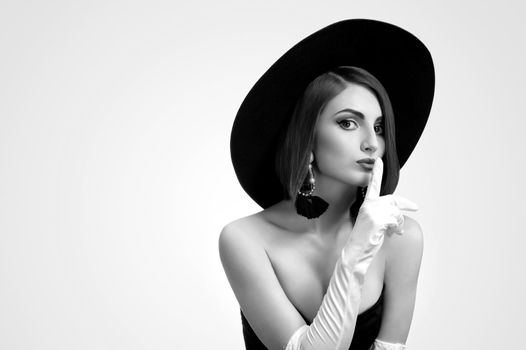 Black and white portrait of a beautiful elegant young fashion model shushing to the camera wearing a hat and gloves posing in a black dress copyspace fashionable stylish luxury beauty concept.