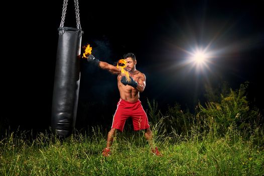 Shirtless muscular male boxer training outdoors at night exercising on a punching bag with his boxing gloves burning with fire copyspace fitness lifestyle forceful powerful confidence athlete.