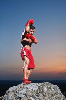 Full length shot of a handsome young male boxer wearing boxing gloves training outdoors sunset on the background fit toned body ripped muscles power masculinity lifestyle champion preparation concept .