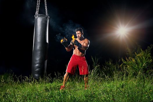 Powerful strong young muscular shirtless boxer with burning gloves training outdoors on a punching bag copyspace fire flames fiery force strength motivation sports sportsperson active athletics.