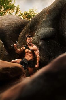 Natural protector. Shot of a fierce powerful young man with muscular body holding his sword looking around at the woods standing on a rock