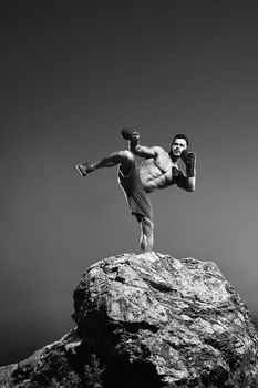 Black and white portrait of a male fighter training outdoors performing kickboxing technique boxing kicking strength balance martial combat fighting masculinity abs fitness sportive athlete physique.