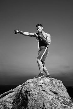 Vertical shot of a handsome young muscular boxer training outdoors on top of a rock confidence determination motivation energy lifestyle sportsman health body power effort endurance combat monochrome.