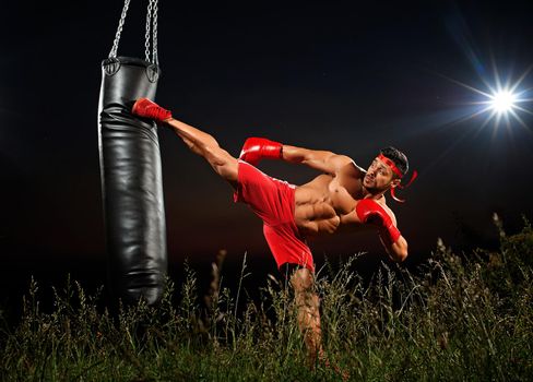 sportsman is training on a black night background, boxer in red shorts and red boxing gloves, muscles, muscular body, night training, training in open space, side kick to a punching bag, black punching bag.