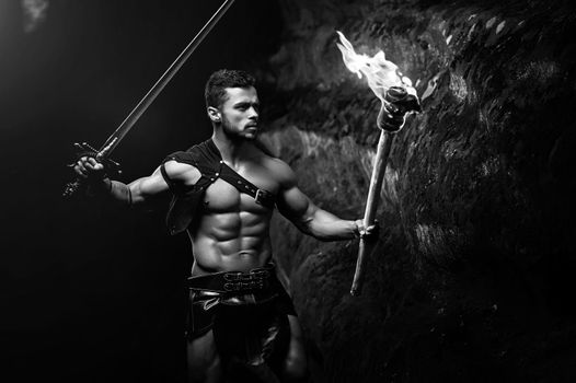 Looking for his path. Monochrome shot of a muscular strong Spartan warrior with a sword walking in the dark forest with a torch looking at the rock