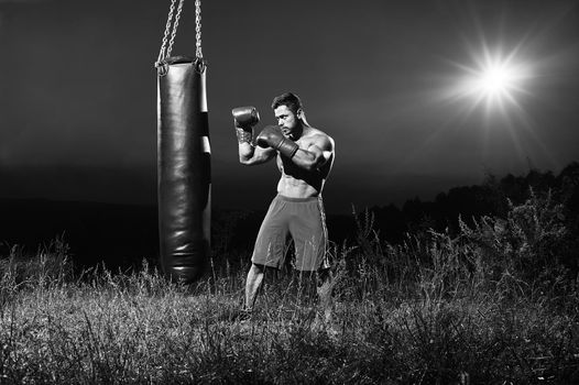 Monochrome portrait of a handsome young muscular male boxer training outdoors at night practicing on a punching bag copyspace nature alone sportsman competitive ambitious strong confident.