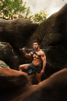 Great fighter he is. Vertical shot of a handsome stunning young man with strong muscular body looking away at the woods holding his sword standing on a rock