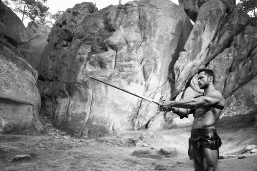 Skillful fighter. Monochrome shot of a warrior with muscular strong body pointing out his sword standing near the rock copyspace