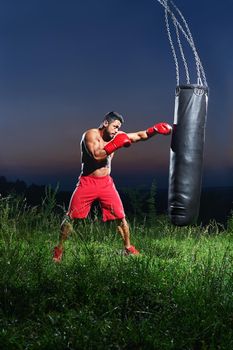 Vertical shot of a shirtless boxer with strong ripped muscular body punching a sandbag training outdoors at night sports motivation lifestyle people sportsman athletics fitness muscles strengthening .