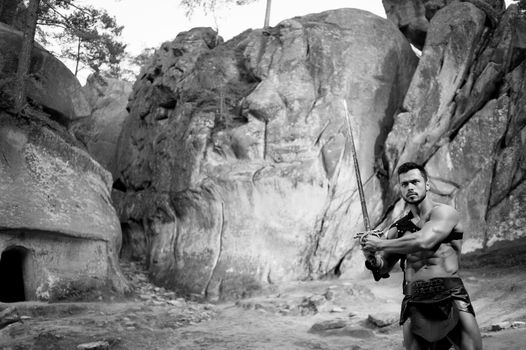 Brave heart. Monochrome portrait of a young warrior with stunning athletic powerful body ready to fight with a sword posing near the rocks