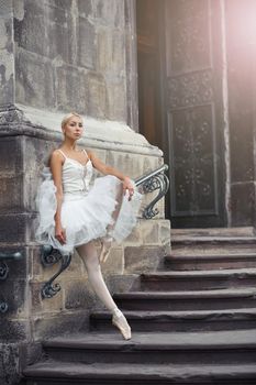 Portrait of a beautiful young blonde ballerina in white outfit standing gracefully on the stairs of an old castle.