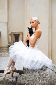 Shot of a beautiful ballerina resting with a black cat in her arms sitting gracefully on the big stone near an old building in the city center.