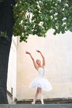 Vertical shot of a ballerina dancing outdoors under the big tree near the building.