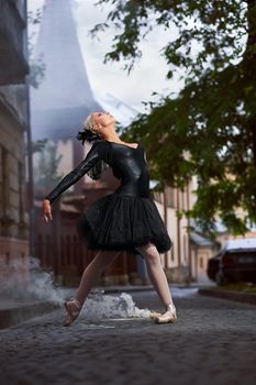 Dramatic dancing performance of a professional ballerina wearing black swan outfit posing gracefully on the smoking street.