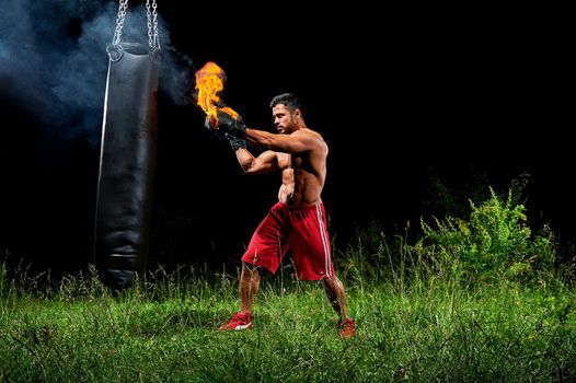 Sport motivation achievement concept. Professional male boxer training on a punching bag outdoors at night wearing burning boxing gloves. Professional martial arts fighter working out on punching bag