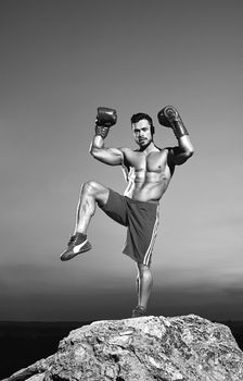 Vertical monochrome shot of a professional male fighter wearing boxing gloves exercising outdoors on top of a rock muscles strength power agility athlete sportsman boxer martial arts combat compete.