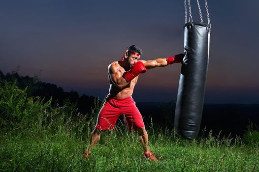 Shirtless muscular young male fighter hitting a punching bag training outdoors on sunset copyspace sportive sports sportsman lifestyle motivation powerful strong muscular abs torso body fitness toning.